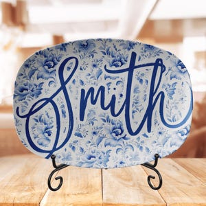 May include: A white oval serving platter with a blue and white floral pattern and the word "Smith" written in blue cursive script. The platter is on a black metal stand.