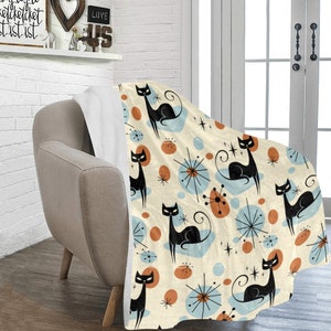 May include: A soft, plush throw blanket with a retro design featuring black cats, orange and blue circles, and starburst shapes on a cream background.