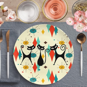 Könnte beinhalten: Ein weißer Keramik-Dinnerteller mit einem Retro-Design mit drei schwarzen Katzen mit stilisierten Schwänzen und einem geometrischen Muster aus roten, gelben, blauen und türkisfarbenen Diamanten. Der Teller hat einen weißen Hintergrund mit einem dezenten Sternenausbruchmuster.