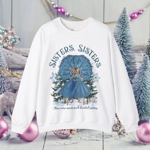 May include: White sweatshirt featuring a blue and white illustration of two sisters standing under a tree with snowflakes. The text "Sisters. Sisters. There were never such devoted sisters" is printed on the sweatshirt.