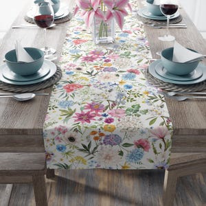 May include: A white table runner with a floral pattern featuring pink, blue, yellow, and green flowers. The runner is on a wooden table with two place settings on each side.