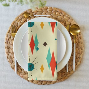 May include: A white dinner plate with a gold rim and a white napkin with a geometric pattern of red, blue, and yellow diamonds and stars on a beige background. The plate and napkin are on a woven wicker placemat.