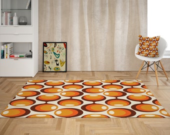 May include: A retro-style rug with a pattern of overlapping orange and brown circles on a cream background. The rug is in a living room setting with a white chair and a framed print on the wall.