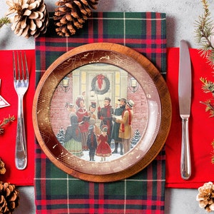 May include: A Christmas-themed dinner table setting with a red and green plaid tablecloth, a wooden plate with a vintage illustration of carolers, a red napkin, a silver fork and knife, and pine branches with pine cones.