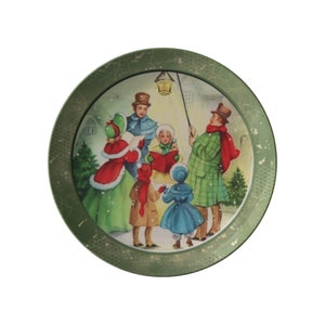 May include: A green ceramic plate with a raised rim features a vintage Christmas scene of people singing carols around a lamppost. The scene is set in a snowy town with houses in the background.