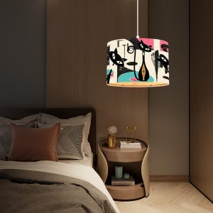 May include: A white pendant light with a black and white cat design on the shade. The shade is cylindrical and has a pink, blue, and green abstract design. The light is hanging over a bed with a brown headboard and white bedding.