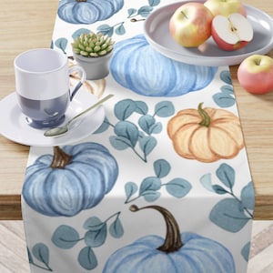 May include: A white table runner with a blue and orange pumpkin pattern. The pumpkins are painted in a watercolor style and are surrounded by green leaves.