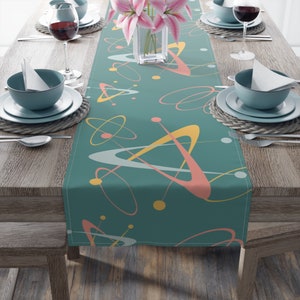 May include: A teal table runner with a mid-century modern atomic design in shades of pink, yellow, and white. The runner is laid out on a wooden table with place settings.