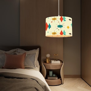 May include: A white pendant light with a colourful geometric pattern on the shade. The shade has a mid-century modern design with red, blue, yellow, and white colours. The light is hanging over a bed with a brown headboard and white bedding.
