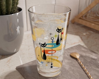 May include: A tall glass with a retro mid-century modern design featuring two black cats and abstract shapes in shades of blue, yellow, orange, and green. The glass is filled with ice and a lemon wedge.