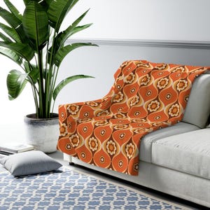 May include: A patterned throw blanket in shades of orange, brown, and cream, draped over a white sofa. The blanket's design features a repeating floral and teardrop motif. A large houseplant in a pot is in the background.