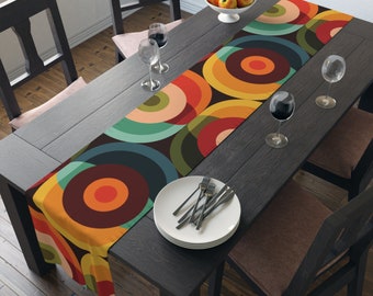 May include: A colorful table runner with a geometric pattern of overlapping circles in shades of orange, yellow, green, blue, and brown. The runner is laid on a dark wood table with two chairs and a bowl of fruit.