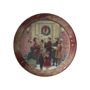 May include: A vintage-style Christmas plate with a red border and a gold-toned inner frame. The plate features a scene of people singing carols in front of a house decorated with a wreath.