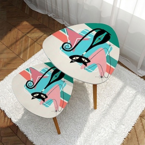 May include: Two retro-style side tables with a mid-century modern design. Each table features a cream-colored surface with a teal, pink, and black cat-like graphic. The tables have three wooden legs and are placed on a white rug.