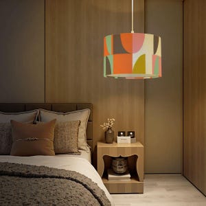 May include: A cylindrical pendant lamp with a colorful geometric pattern of orange, pink, green, and brown shapes. The lamp hangs from a white cord in a bedroom setting. The room has wooden paneling and a bed with pillows and a blanket.