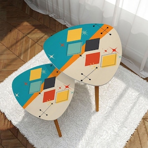 May include: Two retro-style side tables with a mid-century modern design. The tables have a cream-colored surface with a teal blue border and geometric shapes in yellow, red, and black. The tables are supported by wooden legs.