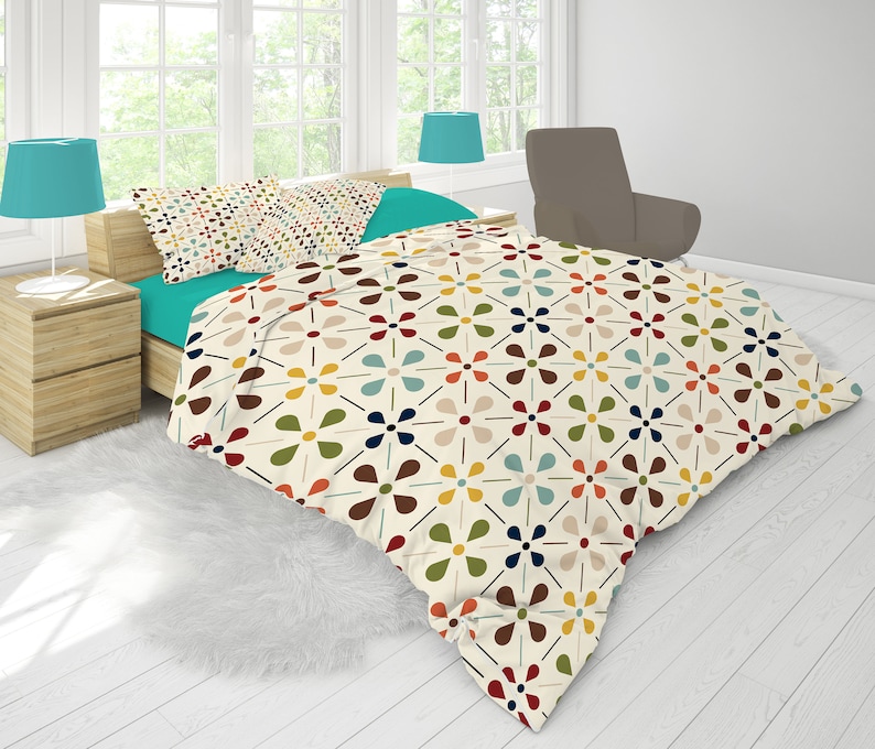 May include: A bed with a cream-colored comforter featuring a repeating floral pattern in red, brown, yellow, blue, and green. The bed has a teal sheet set and matching pillowcases. Two teal lamps sit on wooden nightstands.