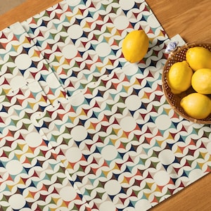 May include: A white table runner with a geometric pattern of overlapping circles and triangles in shades of blue, green, red, yellow, and brown. The runner is folded in half and placed on a wooden table. There are lemons in a wicker basket on the table.