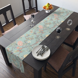 May include: A light blue table runner with a floral and bird design. The runner is on a dark wood table with two chairs and a bowl of fruit.
