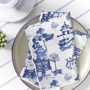 May include: Blue and white patterned fabric napkins featuring a design of Chinese pagodas, trees, and boats. The napkins are folded and displayed on a plate.