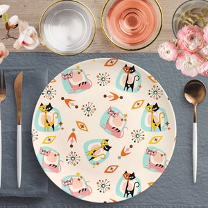 Könnte beinhalten: Ein weißer Keramik-Dinnerteller mit einem Retro-Muster aus bunten Katzen und abstrakten Formen. Der Teller liegt auf einem blauen Platzdeckchen mit einer weißen Serviette und goldenem Besteck.