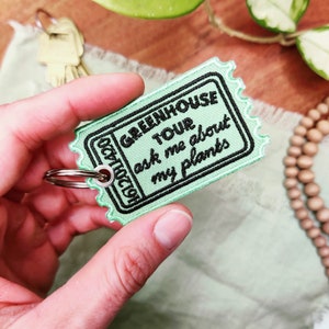 May include: A green ticket-shaped keychain with the text "Greenhouse Tour ask me about my plants" embroidered in black.