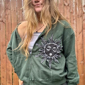 May include: A dark green zip-up hoodie featuring a large, black and white graphic patch of a sun with a human face on the left chest. The hoodie includes a full-length front zipper, long sleeves with ribbed cuffs, and two front pockets.