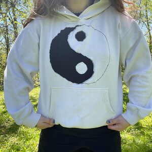 Könnte beinhalten: Cremefarbener Kapuzenpullover mit einem großen schwarz-weißen Yin-Yang-Symbol auf der Vorderseite. Der Hoodie hat eine Vordertasche und eine Kapuze mit Kordelzug. Die Person trägt schwarze Shorts.