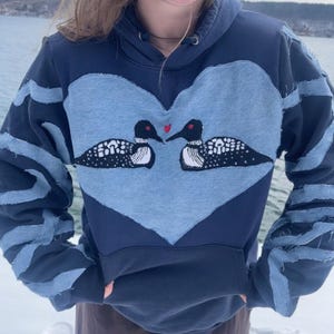 以下が含まれることがあります： ネイビーブルーのパーカー。水色のハート型のパッチには、2羽のハシビロガモのイラストが描かれています。袖とフードには水色のアクセントがあります。ハシビロガモは黒い頭、白い斑点のある体、くちばしの間に赤いハートがあります。