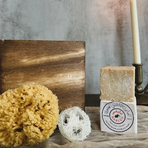 May include: A bar of Nablus olive oil soap, certified organic, with a brown wooden box, a natural loofah sponge, and a white loofah sponge in the background.