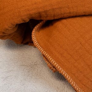 May include: Close-up of a folded, rust-colored fabric with a textured appearance. The edge of the fabric is detailed with a light-colored, stitched border. The fabric is likely linen or a similar material.