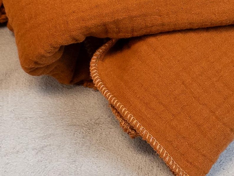 May include: Close-up of a folded, rust-colored fabric with a textured appearance. The edge of the fabric is detailed with a light-colored, stitched border. The fabric is likely linen or a similar material.