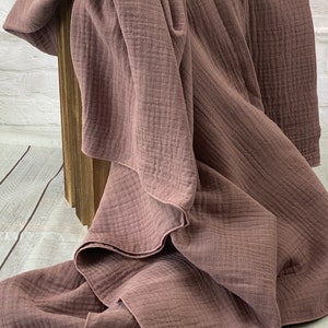 May include: A dusty rose-colored blanket with a textured, crinkled appearance. The blanket is draped over a wooden structure, showcasing its soft, flowing folds. The fabric appears lightweight and breathable, suggesting a cozy textile for home decor.