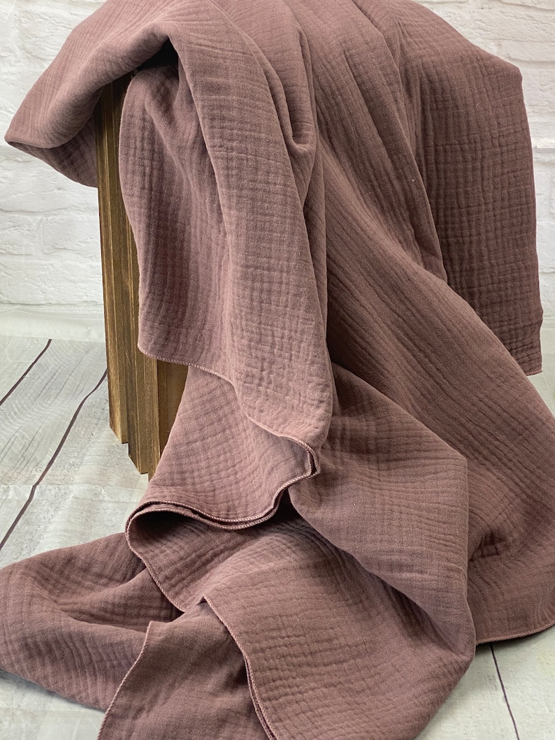 May include: A dusty rose-colored blanket with a textured, crinkled appearance. The blanket is draped over a wooden structure, showcasing its soft, flowing folds. The fabric appears lightweight and breathable, suggesting a cozy textile for home decor.