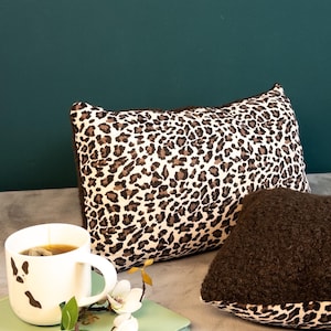Könnte beinhalten: Eine weiße Tasse mit schwarzen Kuhflecken steht neben einem Kissen mit Leopardenmuster und einem braunen, strukturierten Kissen. Ein Zweig mit weißen Blumen und ein grünes Notizbuch sind ebenfalls im Bild. Der Hintergrund ist dunkelgrün.