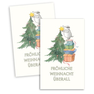 Könnte beinhalten: Zwei Weihnachtskarten mit einer Aquarellillustration. Ein Dachs steht auf einem Geschenkeberg und setzt einen Stern auf einen Weihnachtsbaum. Der Baum ist mit roten Kugeln geschmückt. Der Text "FRÖHLICHE WEIHNACHT ÜBERALL" ist darunter gedruckt.