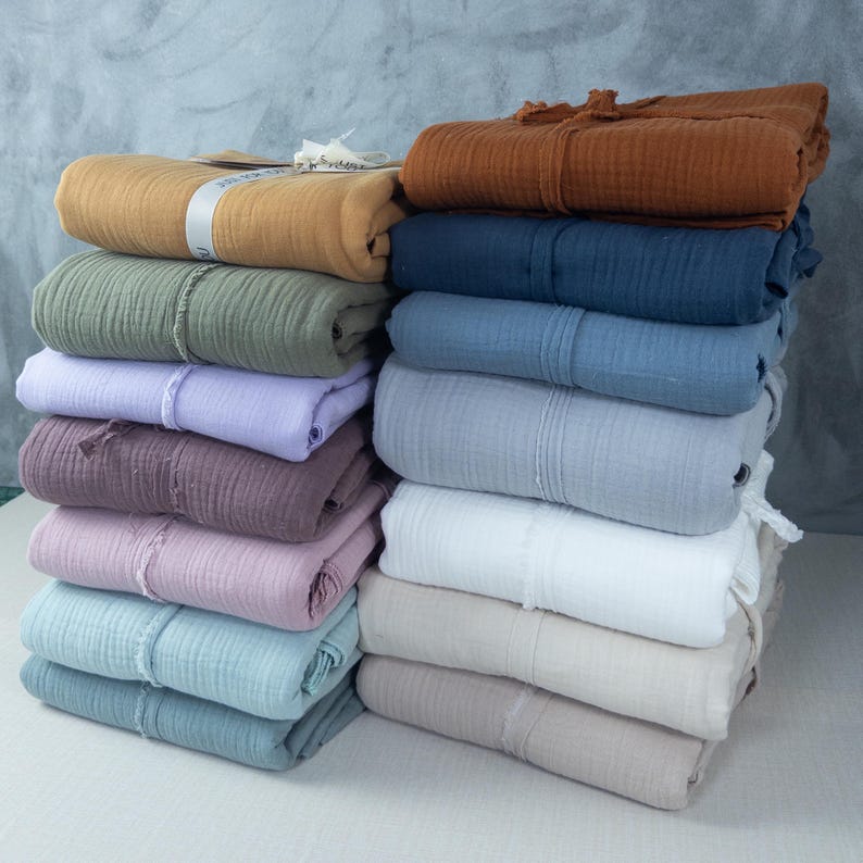 May include: A stack of folded, soft-looking fabric in various colors, including mustard yellow, olive green, lavender, and dusty rose. The fabric appears to be a lightweight, textured material, possibly gauze or muslin. Each piece is neatly rolled and tied.