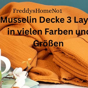 May include: An orange-colored muslin blanket, folded and stacked, with the text "FreddysHomeNo1" and "Musselin Decke 3 Layer in vielen Farben und Gr&ouml;&szlig;en". A white mug and floral arrangement are in the foreground.