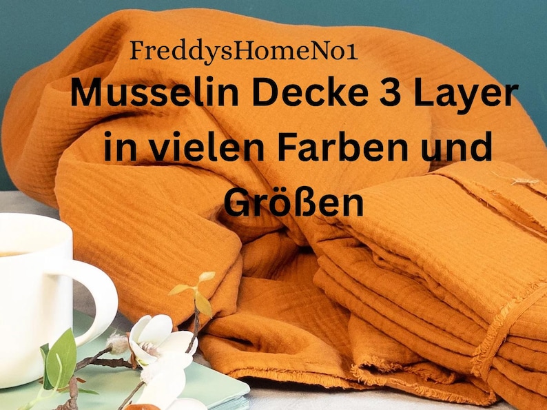 May include: An orange-colored muslin blanket, folded and stacked, with the text "FreddysHomeNo1" and "Musselin Decke 3 Layer in vielen Farben und Gr&ouml;&szlig;en". A white mug and floral arrangement are in the foreground.