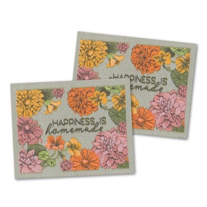 Könnte beinhalten: Zwei rechteckige Stoffaufnäher mit floraler Stickerei. Die Aufnäher zeigen orange, rosa und gelbe Blumen mit grünen Blättern. Der Text "HAPPINESS IS homemade" ist in Schwarz gestickt.