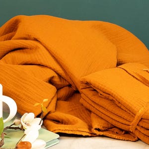 May include: Mustard-colored muslin fabric, artfully draped and folded, with a stack of folded fabric tied with a matching ribbon. A white mug and floral arrangement are in the foreground, suggesting a cozy, homey aesthetic.