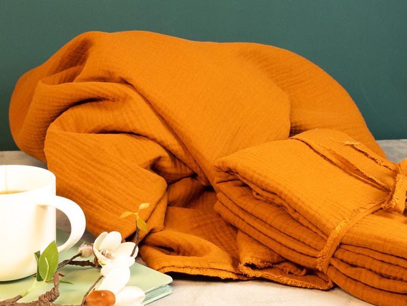 May include: Mustard-colored muslin fabric, artfully draped and folded, with a stack of folded fabric tied with a matching ribbon. A white mug and floral arrangement are in the foreground, suggesting a cozy, homey aesthetic.