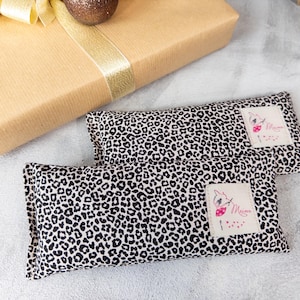 Pode incluir: Duas almofadas retangulares com um padrão de leopardo em preto e branco. Cada almofada tem uma pequena etiqueta com um gráfico rosa e branco e a palavra "Maime". Um presente embrulhado com uma fita dourada e um adorno dourado no fundo.