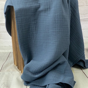 May include: A folded, dark blue muslin blanket. The blanket is draped over a wooden box, with the fabric's texture and color prominently displayed. The blanket's edges are visible, showcasing its soft, flowing nature.