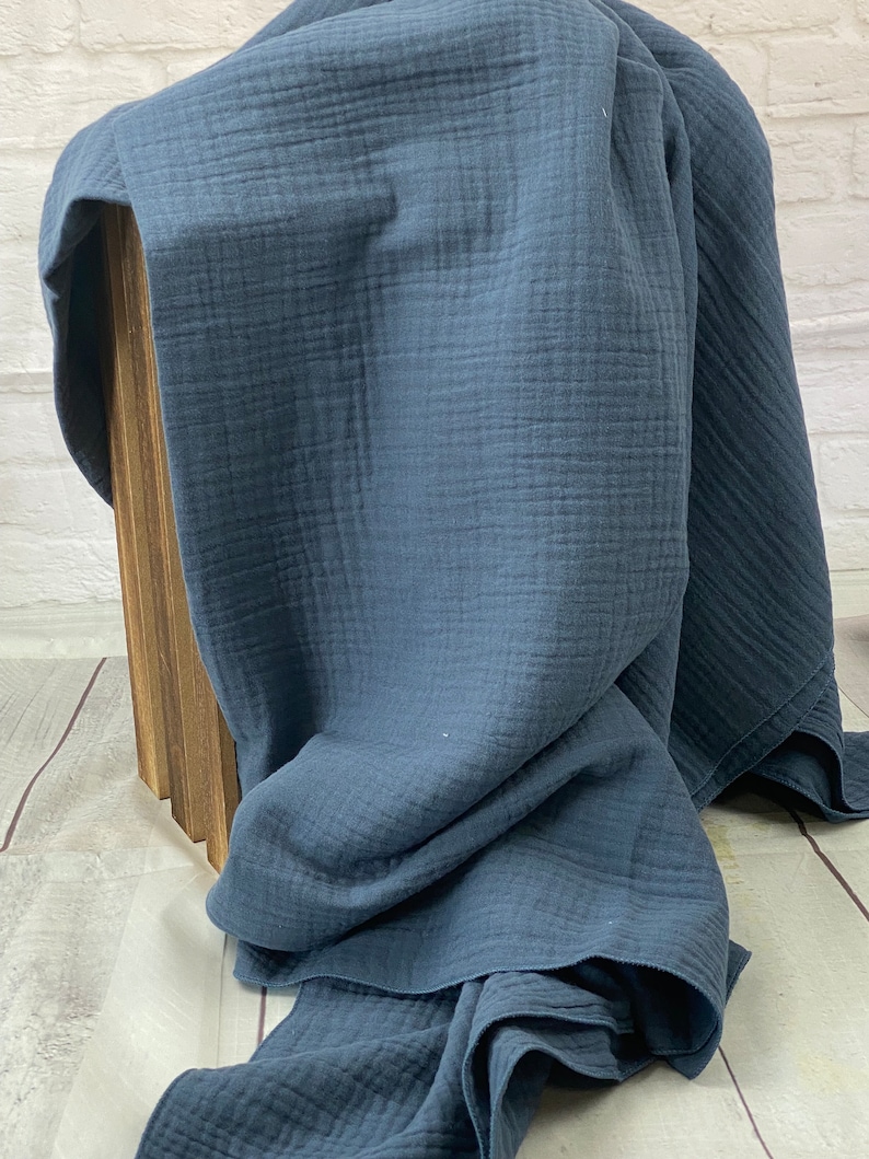 May include: A folded, dark blue muslin blanket. The blanket is draped over a wooden box, with the fabric's texture and color prominently displayed. The blanket's edges are visible, showcasing its soft, flowing nature.