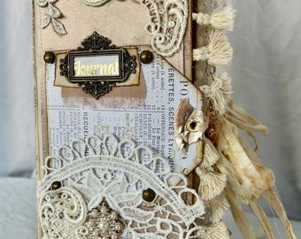Puede incluir: Un diario hecho a mano con una estética vintage. La cubierta beige está adornada con encaje blanco, detalles de perlas y una etiqueta metálica que dice "Journal". El lomo presenta borlas y cintas color crema. El diario está decorado con efímeras vintage.