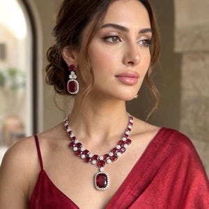 May include: A woman wearing a ruby and diamond jewellery set. The set includes a necklace with a large pendant, matching earrings, and a red dress. The jewellery features a combination of red gemstones and clear diamonds.