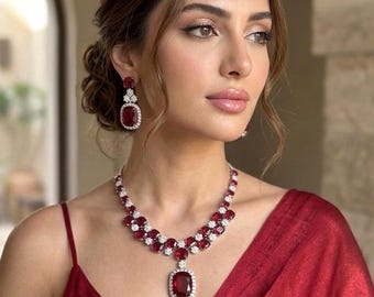 Puede incluir: Una mujer con un conjunto de joyas de rubíes y diamantes. El conjunto incluye un collar con un colgante grande, pendientes a juego y un vestido rojo. Las joyas presentan una combinación de piedras preciosas rojas y diamantes transparentes.
