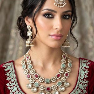 May include: A woman wearing a gold-toned headpiece, earrings, and a layered necklace. The necklace features red and green gemstones set in a floral design. The woman is wearing a red velvet top with embroidered detailing.