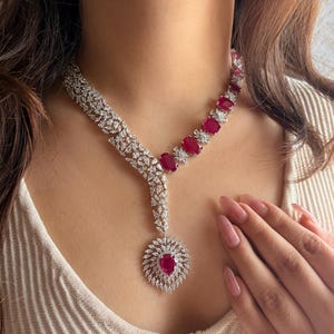 Può includere: Una collana d'argento con un grande ciondolo di pietra rossa e pietre rosse più piccole lungo la catena. La collana è realizzata in argento e presenta un delicato design floreale.